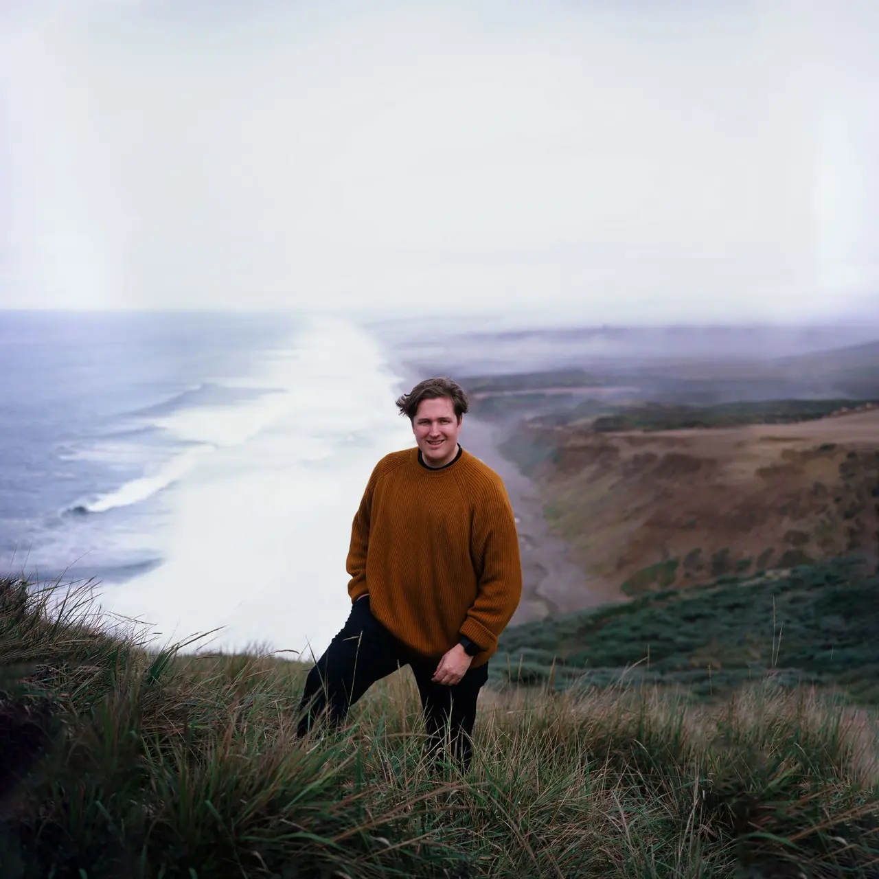 Jake in a rust wool sweater on a grass bluff, the Point Reyes shoreline and fog behind him
